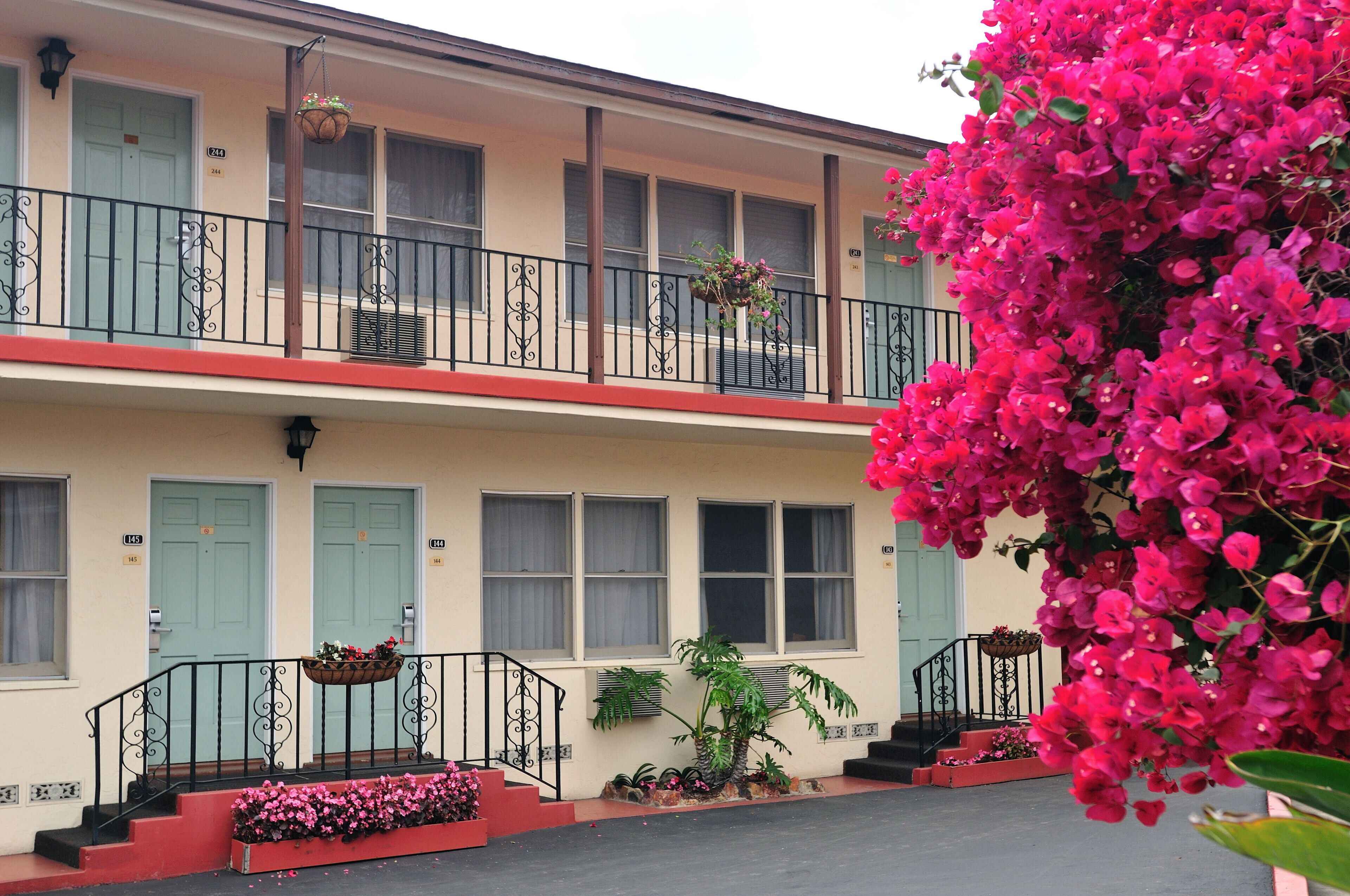 Un edificio de dos pisos con un balcón, un cantero con flores rosas y una planta en maceta.