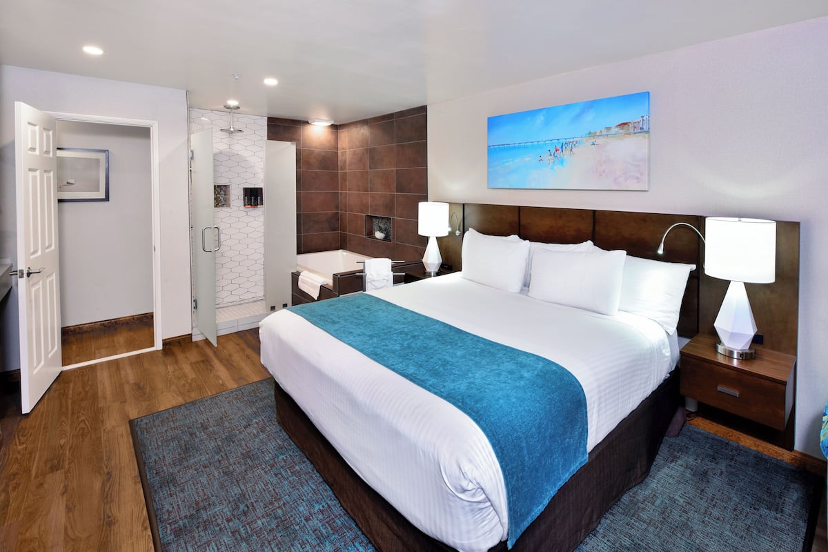 A hotel room with a large bed, a bathroom with a shower, a bedside table with a lamp, and a painting on the wall.