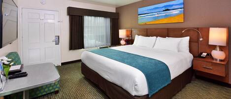 A hotel room with a large bed, a desk, a chair, a window with curtains, a painting on the wall, and a nightstand with a lamp.
