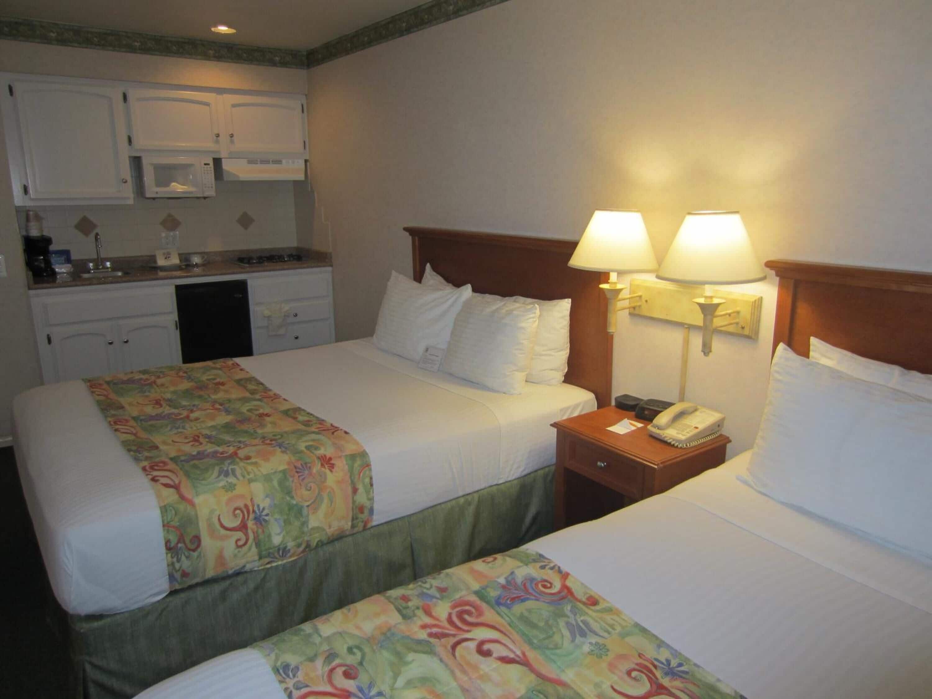 A hotel room with two beds, a small kitchenette, and a bedside lamp.