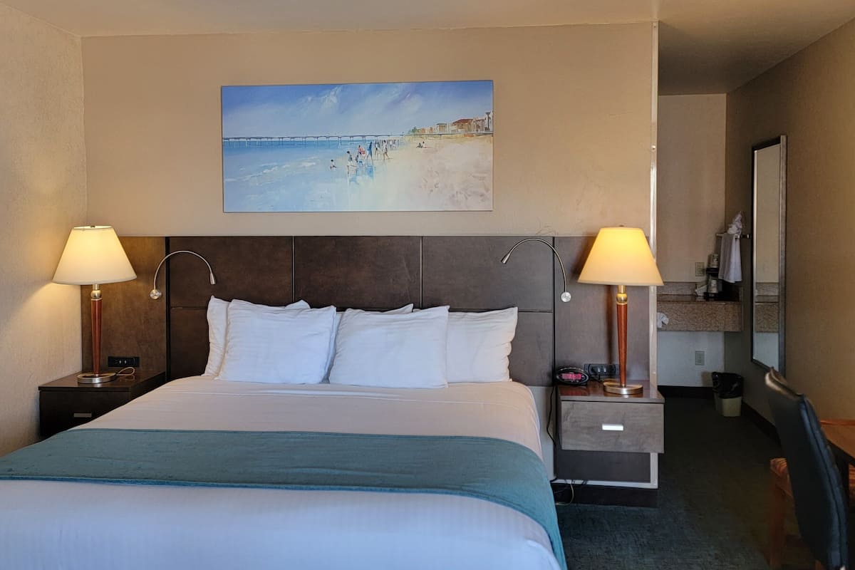 A hotel room with a large bed, two bedside tables with lamps, a painting of a beach scene, and a view of the room's interior.