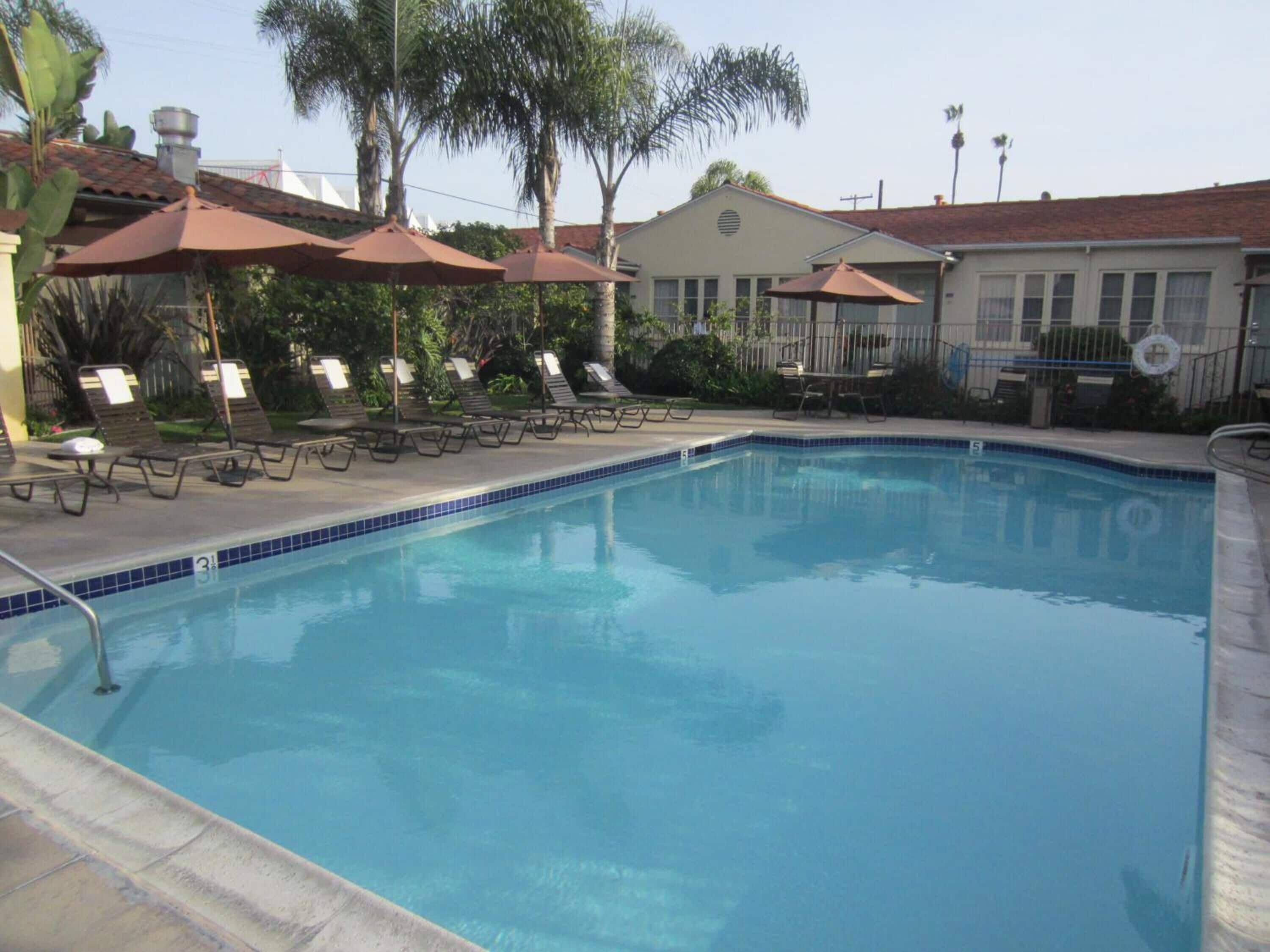 A swimming pool with lounge chairs and umbrellas, surrounded by palm trees and a building with a red roof.
