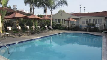 A swimming pool with lounge chairs and umbrellas, surrounded by palm trees and a building with a red roof.
