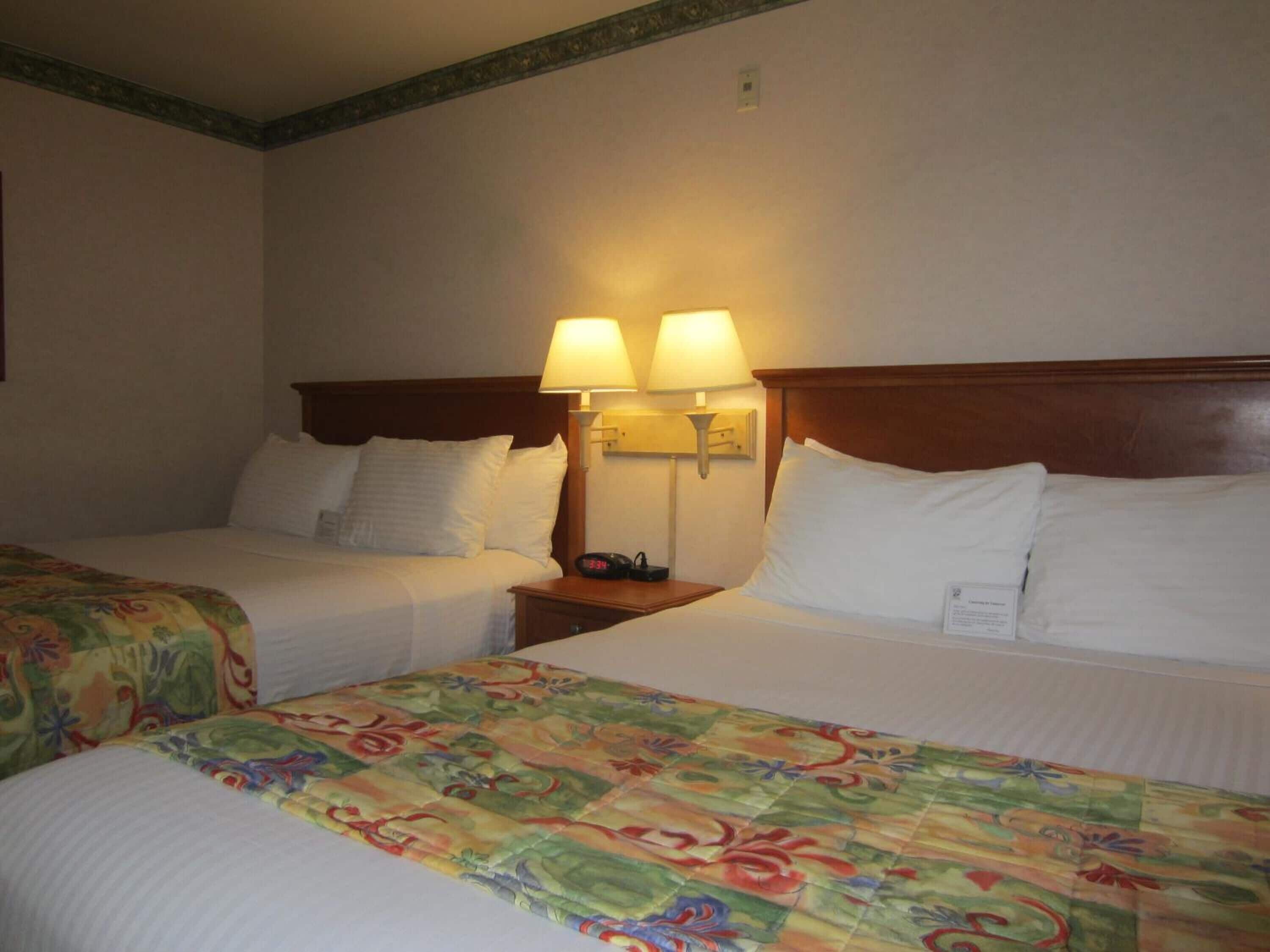 A hotel room with two beds, each with white linens and a floral bedspread. There are two wall-mounted lamps providing light.
