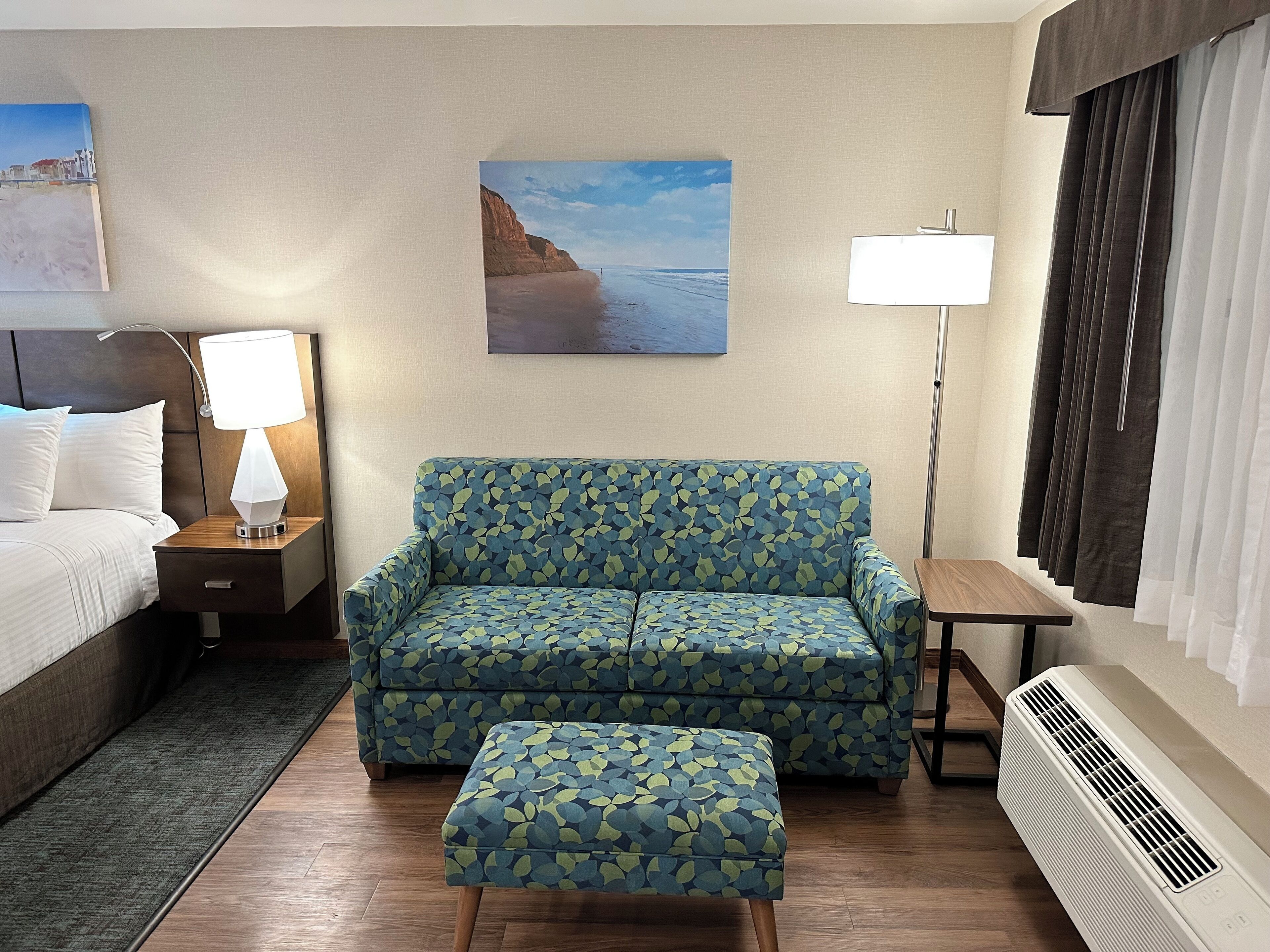 A hotel room with a bed, a sofa, an ottoman, a nightstand with a lamp, a painting of a beach, a standing lamp, and a window with curtains.