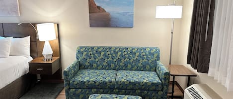 A hotel room with a bed, a sofa, an ottoman, a nightstand with a lamp, a painting of a beach, a standing lamp, and a window with curtains.