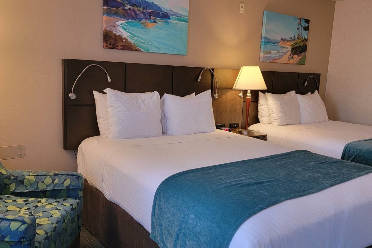 A hotel room with two beds, a nightstand, a lamp, and artwork on the walls.