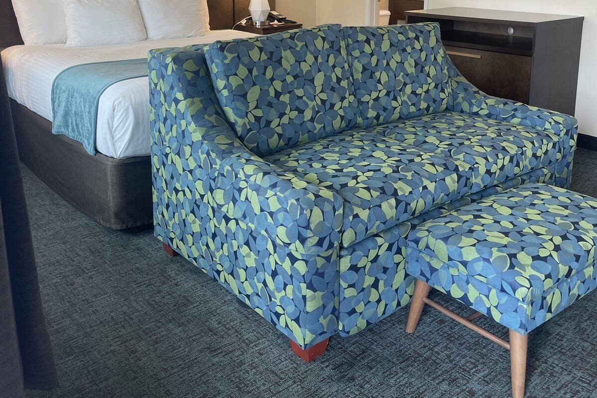 A hotel room with a bed, a sofa, a desk, a ceiling fan, a painting, and a framed picture on the wall.
