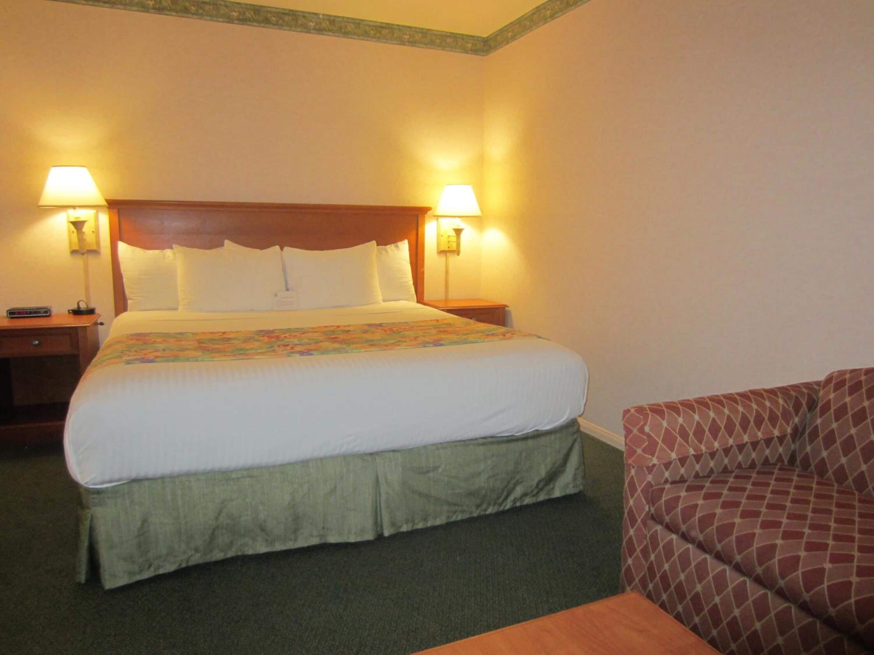 Una habitación de hotel con una cama grande, dos lámparas de noche, una mesita pequeña y un sillón estampado.
