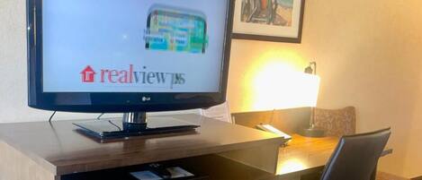 A hotel room with a large flat-screen TV displaying 'realview ps' on the screen, a desk with a lamp, a chair, and a small drawer unit.