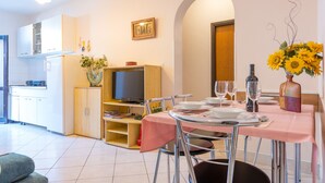 Uma sala de jantar com uma mesa posta para quatro pessoas, uma televisão e uma cozinha ao fundo.