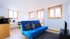 Una sala de estar moderna con un sofá azul, un escritorio pequeño con un televisor, y una cocina al fondo.