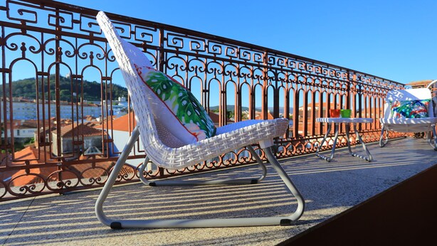 Una silla de mimbre blanca con un cojín colorido, ubicada en un balcón con barandilla ornamental de hierro forjado.