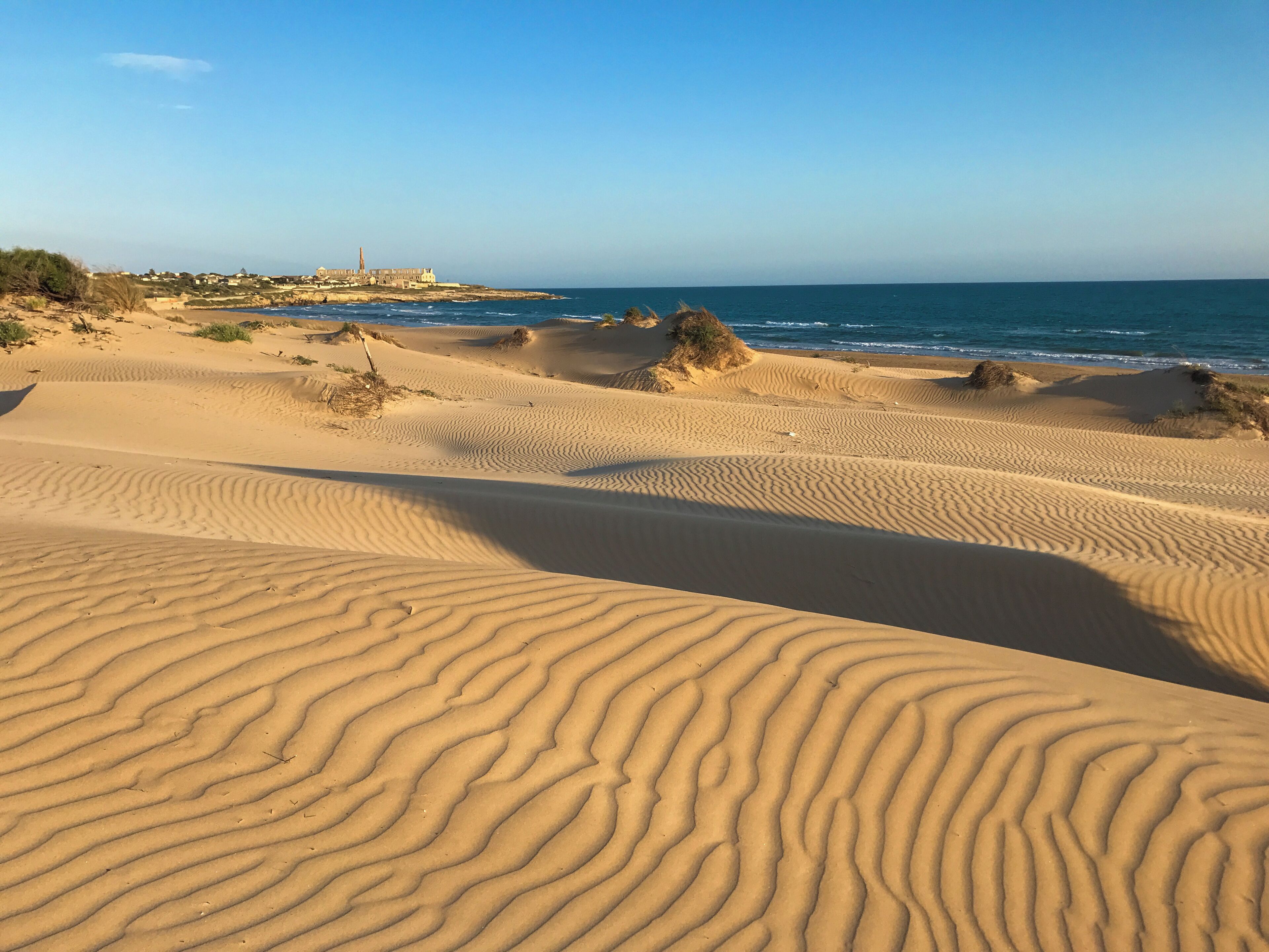 A desert landscape with sand dunes and a distant view of a coastal town.