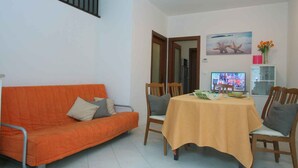 Ein Wohnzimmer mit einem orangefarbenen Sofa, Holzstühlen, einem Esstisch mit gelber Tischdecke, einem Fernseher und einem Gemälde von Meereslebewesen.