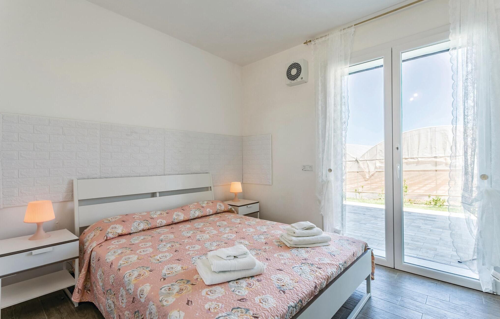 A bedroom with a white brick-patterned headboard, a floral bedspread, two bedside lamps, a window with sheer curtains, and a view of a garden outside.