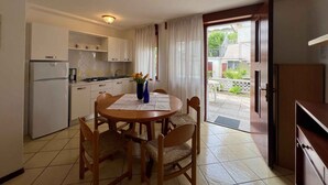 A compact kitchen with white cabinets, a refrigerator, and a dining table with chairs. A window with curtains is visible, and a door leads to an outdoor area. - Holiday apartment with open terrace and air conditioning (Lignano)
