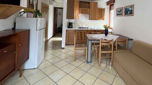 A compact kitchen with wooden cabinets, a dining table with chairs, a refrigerator, and a TV mounted on the wall. - Holiday apartment with open terrace and air conditioning (Lignano)