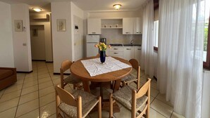 A dining area with a round wooden table, four wicker chairs, a vase with flowers, and a kitchen in the background. - Holiday apartment with open terrace and air conditioning (Lignano)