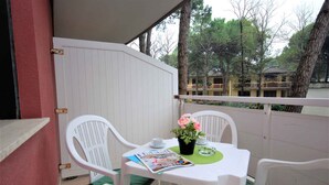 A balcony with white chairs and a table, a magazine, a potted plant, and a view of trees and buildings. - Holiday apartment with balcony and pool (Lignano)