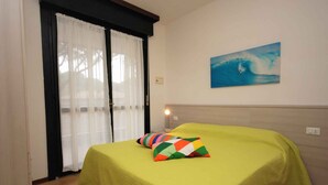 A room with a bed covered in a green bedspread, a colorful pillow, a painting of a wave on the wall, and a window with white curtains. - Holiday apartment with balcony and pool (Lignano)