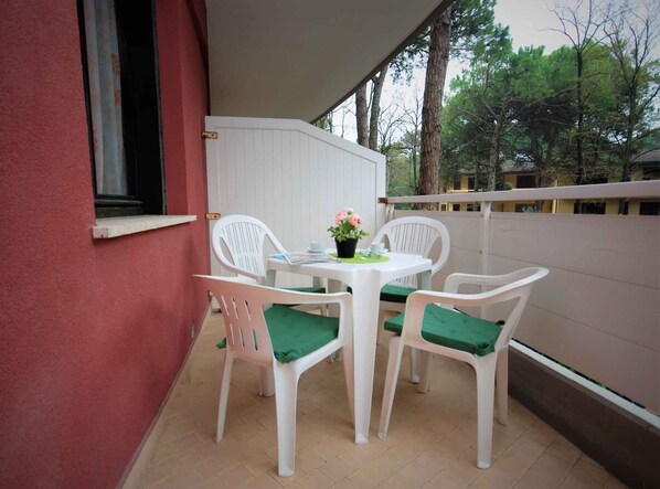 A small patio with a white table and four white chairs, each with a green cushion. There is a window on the left and a white fence in the background.