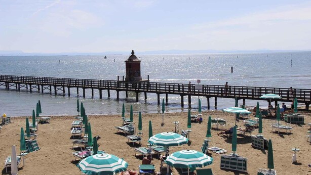 Une plage bordée de rangées de chaises longues et de parasols, une jetée qui s’avance dans l’eau et un phare visible au loin.