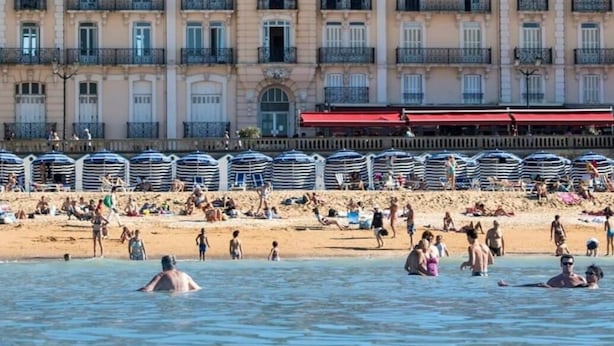 Een strand met mensen die zwemmen en zonnebaden, een rij strandparasols, en een gebouw met balkons op de achtergrond.