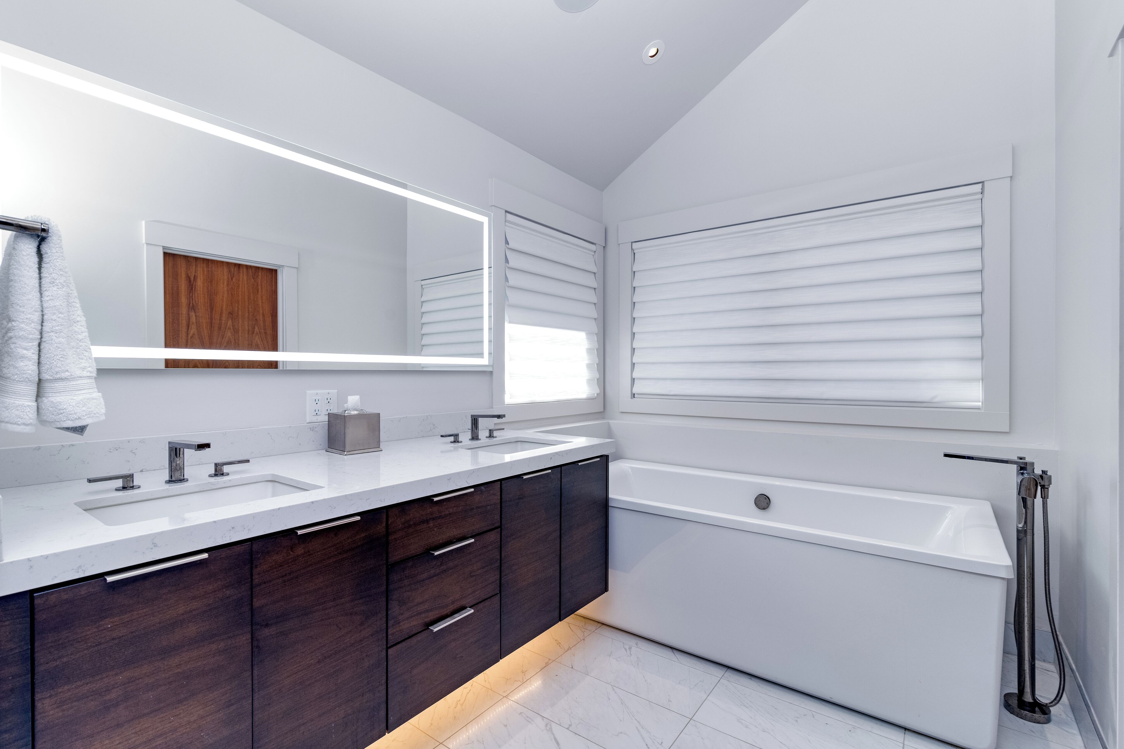 Un baño moderno con un espejo grande, lavamanos dobles, armarios de madera oscura, una bañera blanca y una ventana con persianas.