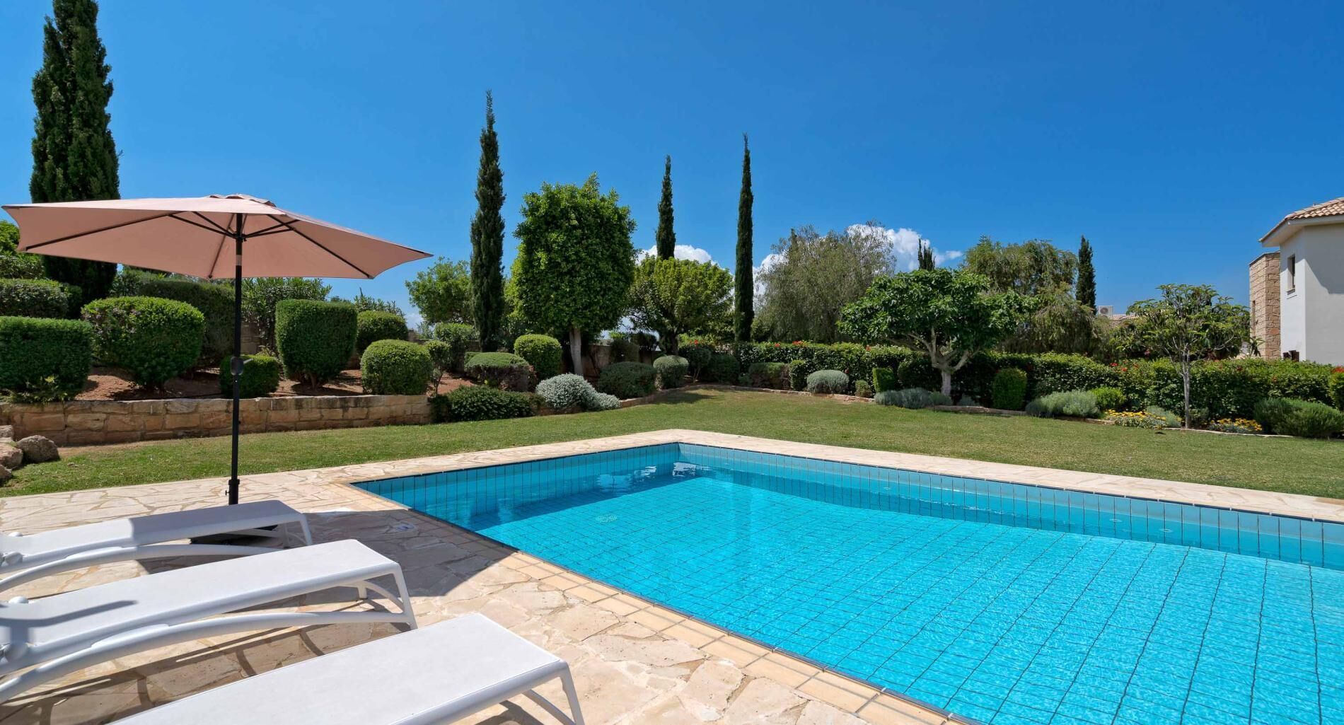 A rectangular swimming pool with a tiled edge, surrounded by a stone patio. There are white lounge chairs and a single umbrella.