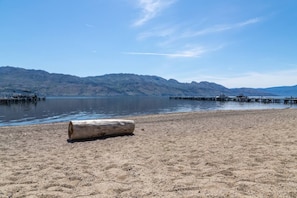 Une plage de sable avec un tronc d’arbre au premier plan, une jetée s’étendant dans une eau calme, et des montagnes en arrière-plan.
