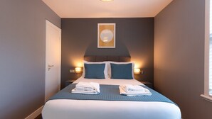 Une chambre d’hôtel avec un lit, deux oreillers, une table de chevet, un luminaire fixé au mur et un tableau encadré accroché au mur.
