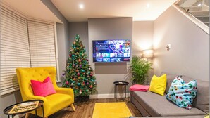 Un salon avec un fauteuil jaune, un canapé gris, un sapin de Noël et un téléviseur à écran plat fixé au mur.