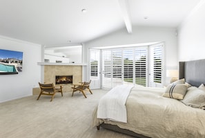 Une chambre à coucher avec un grand lit, une cheminée, un coin salon avec des fauteuils et une vue sur l’extérieur, visible à travers une porte vitrée.