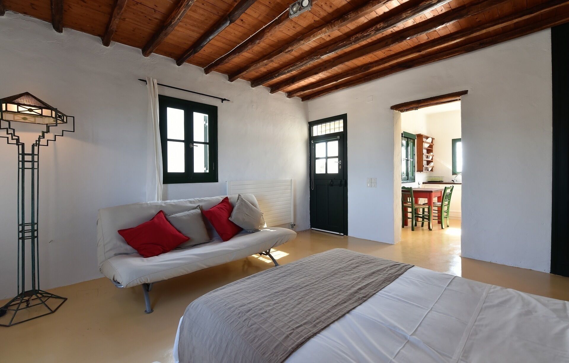 A room with a white sofa, a bed, a floor lamp, and a door leading to a kitchen area.
