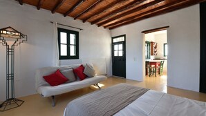 A room with a white sofa, a bed, a floor lamp, and a door leading to a kitchen area. - Santa Maria Studio 5 Mins From the Beach (Paros)