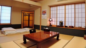 Une pièce de style japonais traditionnel, avec un sol recouvert de tatamis, une petite table en bois, un futon servant de lit, un paravent coulissant de type shoji et une petite étagère où sont posés des objets personnels.