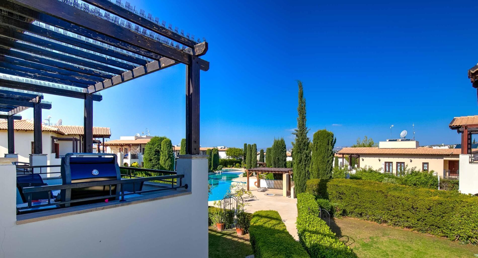 A poolside area with a pergola, a barbecue grill, and a view of a swimming pool and surrounding greenery.
