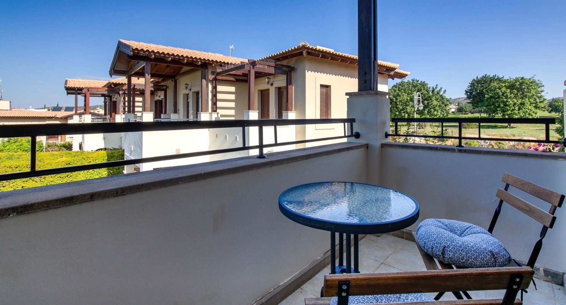A balcony with a round table and a wooden bench, overlooking a building with a tiled roof and a garden.