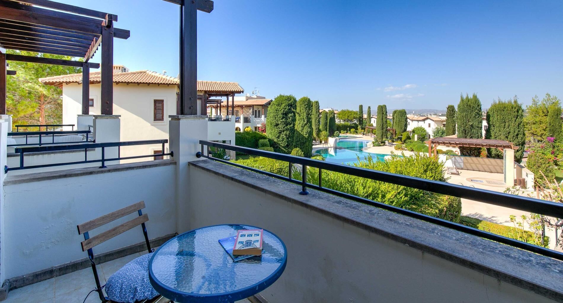 A balcony with a glass table, a wooden bench, and a view of a swimming pool and surrounding buildings.