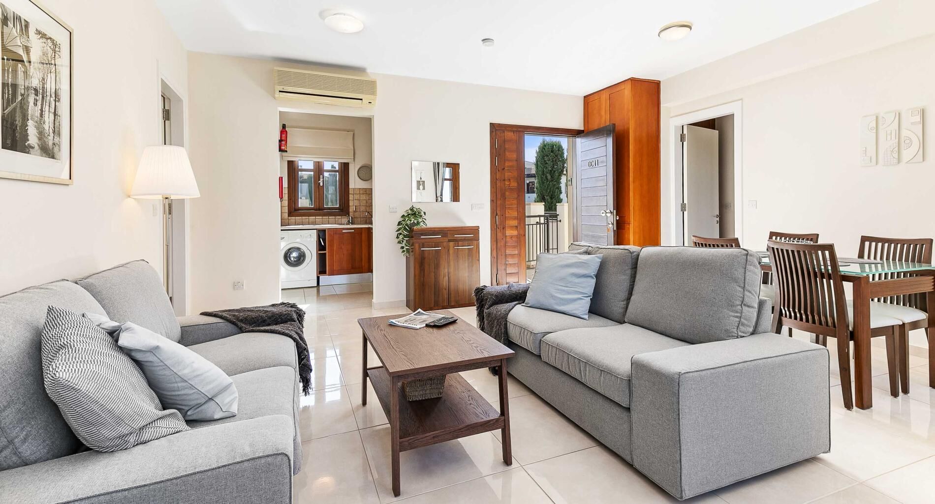 A modern living room with grey sofas, a wooden coffee table, and a dining area with wooden chairs and a glass table.
