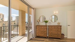 A balcony with a view of buildings, a wooden dresser with drawers, a round mirror, and a clock on the wall. - Waikiki Moana Inn (Honolulu)