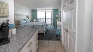 A hotel room with a bed, a small desk, a coffee maker, a window with curtains, and a door with a towel rack. - Sand Dunes 2434 (Myrtle Beach)