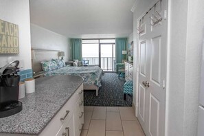 A hotel room with a bed, a small desk, a coffee maker, a window with curtains, and a door with a towel rack.