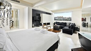 Ein modernes Hotelzimmer mit einem großen Bett, einer Sitzecke mit Sofa und Sesseln, einem Flachbildfernseher und Blick auf eine Stadtlandschaft.