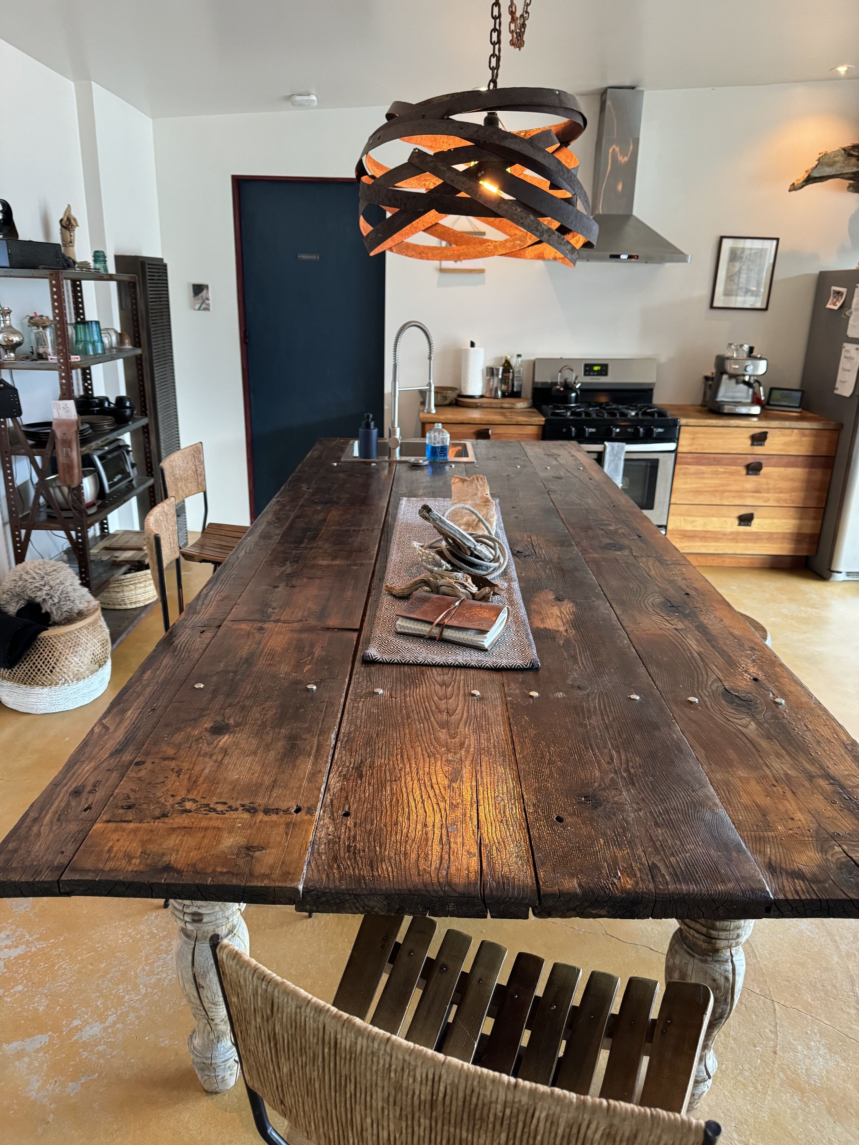 A rustic wooden dining table with a metal chandelier, surrounded by chairs and a kitchen in the background.