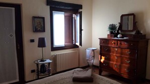 Un dormitorio con cama, cómoda, ventana con cortinas, radiador y una mesita de noche con lámpara.
