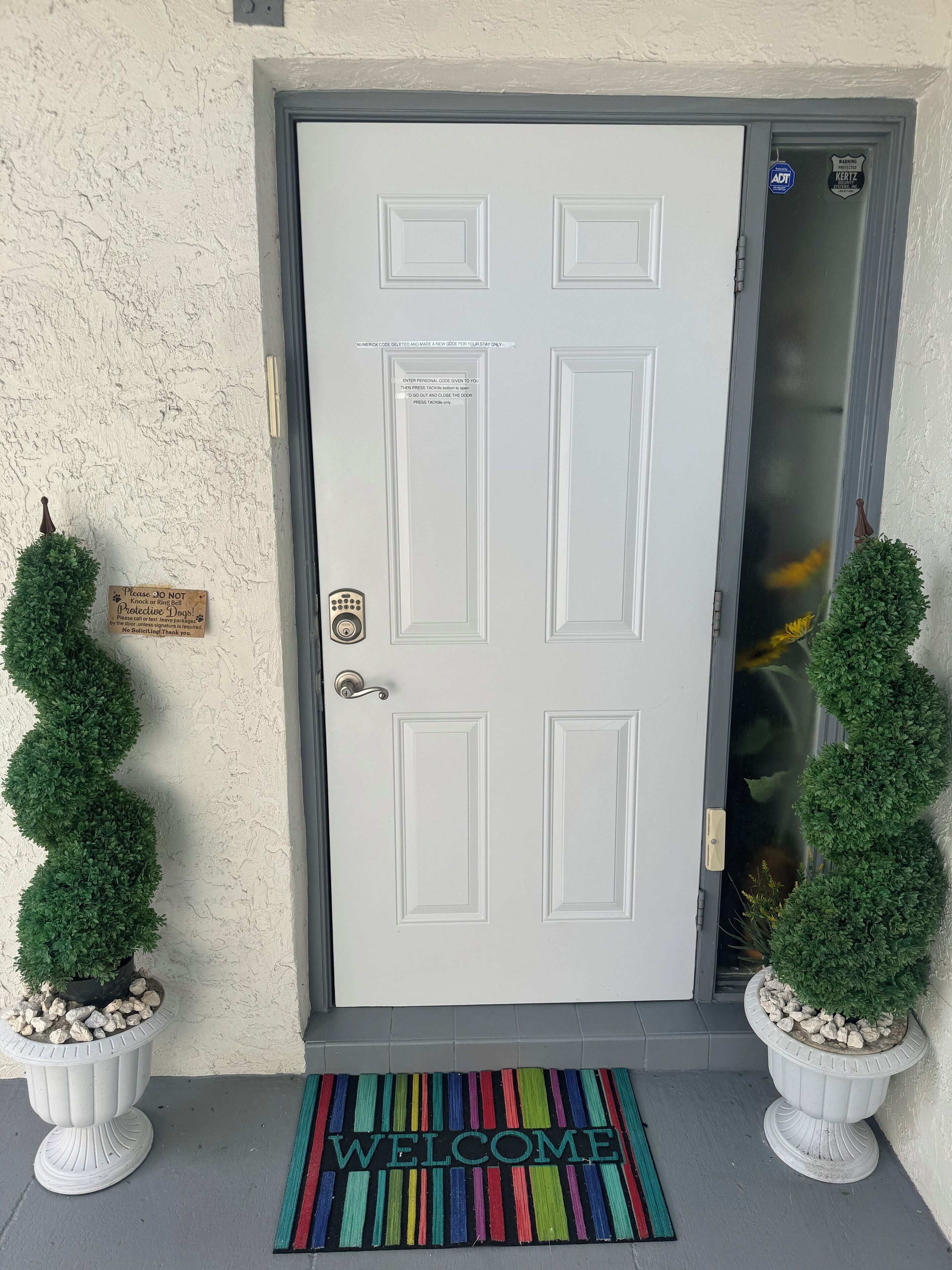 Une porte blanche dotée d’un système de verrouillage à code, un paillasson avec l’inscription « Bienvenue » et deux plantes en pot, une de chaque côté.
