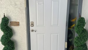 Una puerta blanca con cerradura de código de seguridad, un tapete de 'Welcome' y dos macetas con plantas a cada lado.