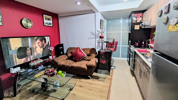 A modern living room with a brown sofa, a glass coffee table, and a flat-screen TV. A kitchen with a refrigerator and stove is visible in the background. - Complete Apartment in Coapa CDMX (Ciudad de México)